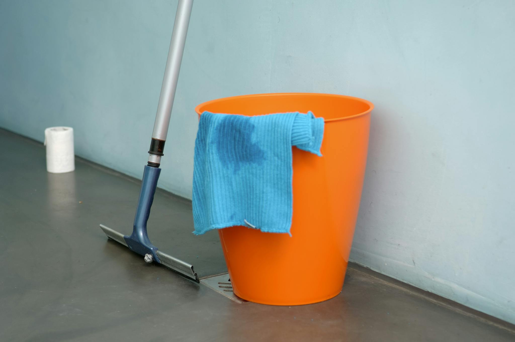 Close-up of cleaning supplies including orange bucket, mop, and blue rag indoors.